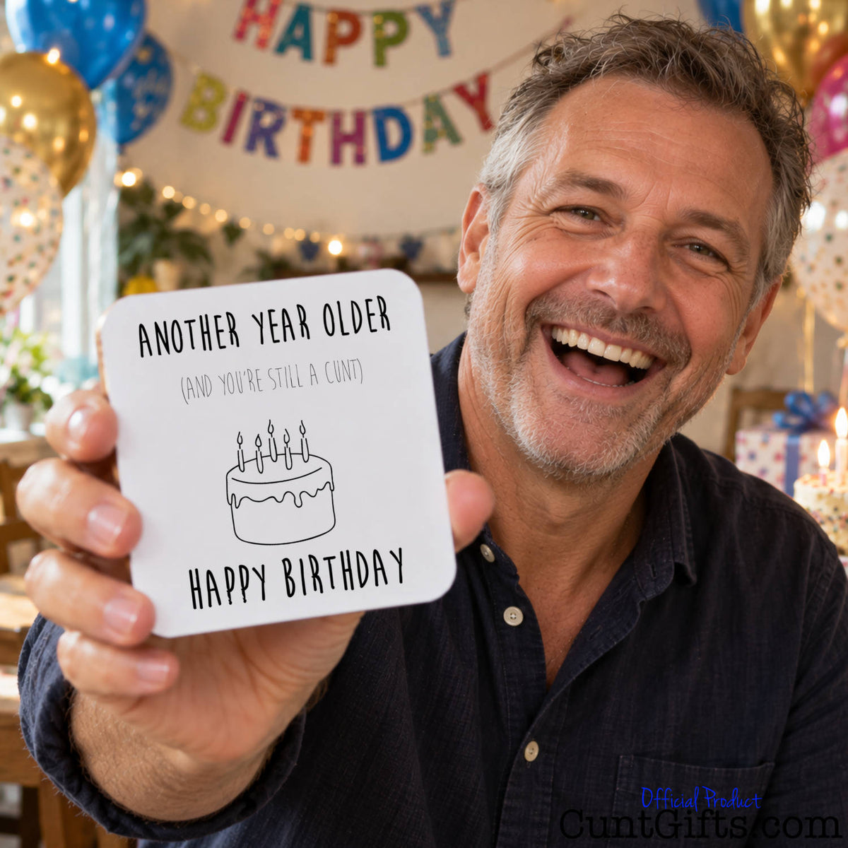 A smiling man in a navy shirt holds up a square white birthday drinks coaster card that reads 'ANOTHER YEAR OLDER (AND YOU'RE STILL A CUNT) HAPPY BIRTHDAY' with a simple cake illustration. The background shows a festive party setting with balloons, lights, and a 'Happy Birthday' banner.