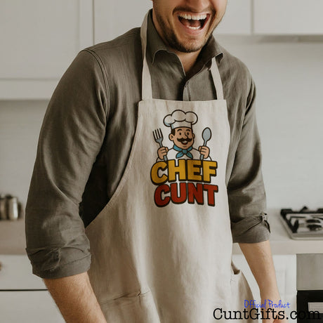 Man wearing a linen Chef Cunt apron in a kitchen, laughing.