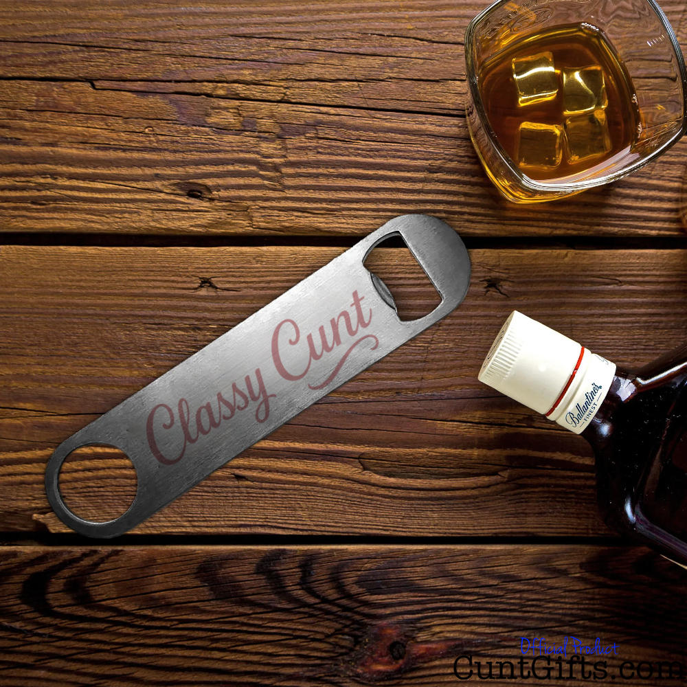 Metal bottle opener with 'Classy Cunt' design on a wooden surface with a glass of whiskey.