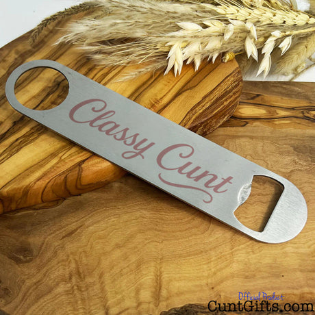 Metal bottle opener with 'Classy Cunt' design on a wooden table with wheat.