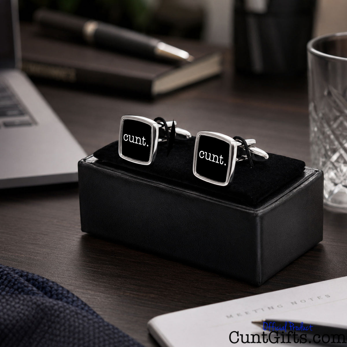 A pair of square silver cufflinks with black faces and the word 'cunt.' in white, displayed on a black presentation box on a dark wooden desk with office items in the background.