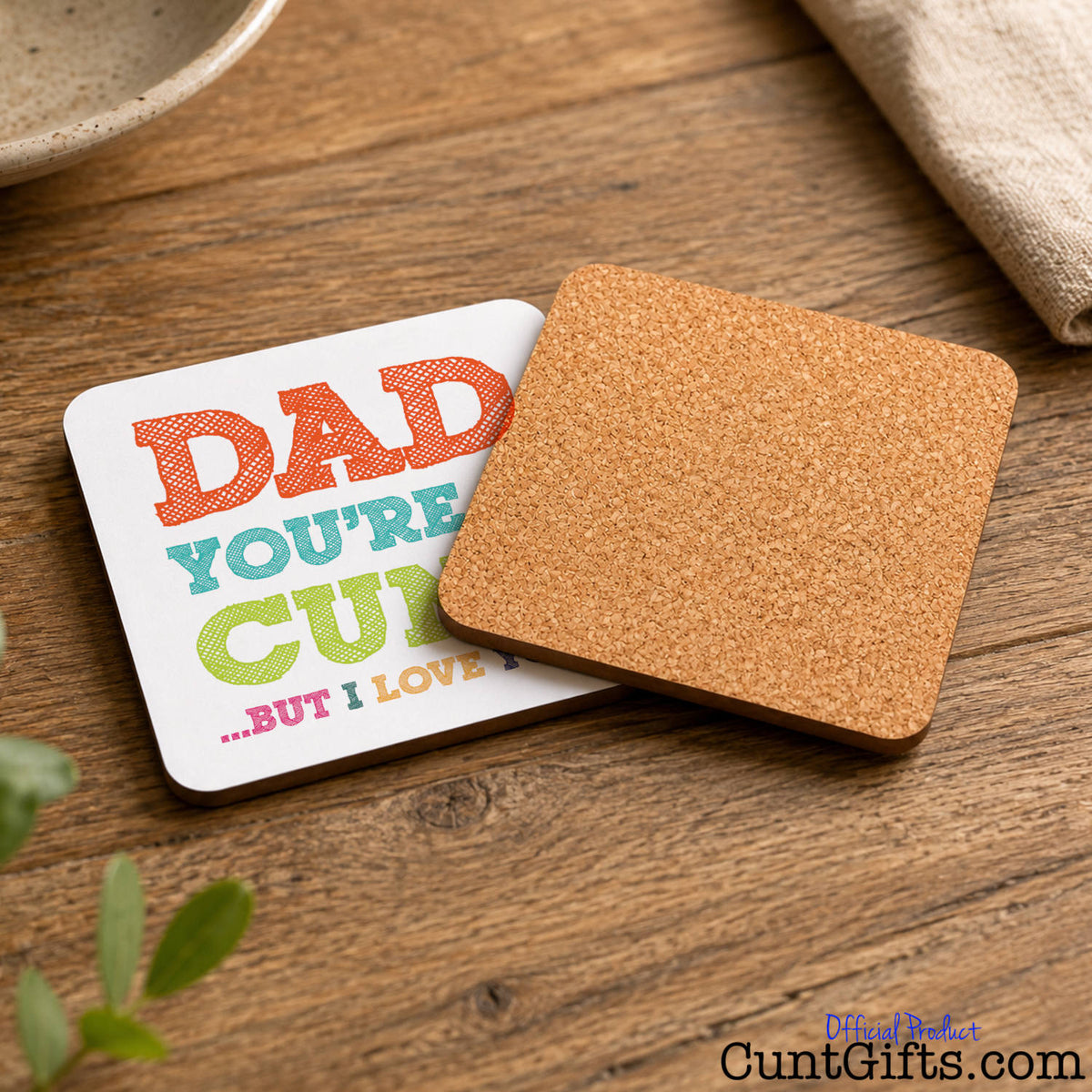 A square cork coaster resting on top of a white printed coaster that reads “Dad You're A Cunt But I Love You".
