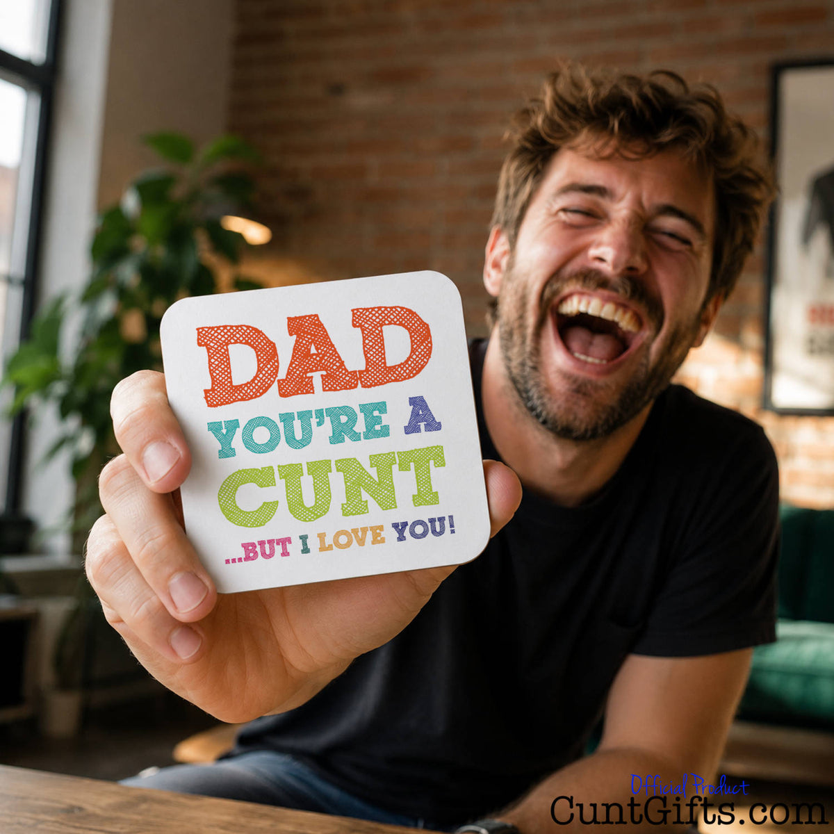 A man in a black t-shirt is laughing while holding up a square white drink coaster with colorful text that reads "Dad You're A Cunt But I Love You".