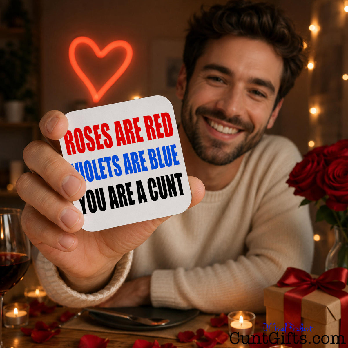 A smiling man at a romantic table setting holds up a white square drinks coaster with the text 'ROSES ARE RED VIOLETS ARE BLUE YOU ARE A CUNT' in red, blue, and black lettering. The background is warmly lit with a glowing red heart neon sign, string lights, red roses, candles, and a gift box.