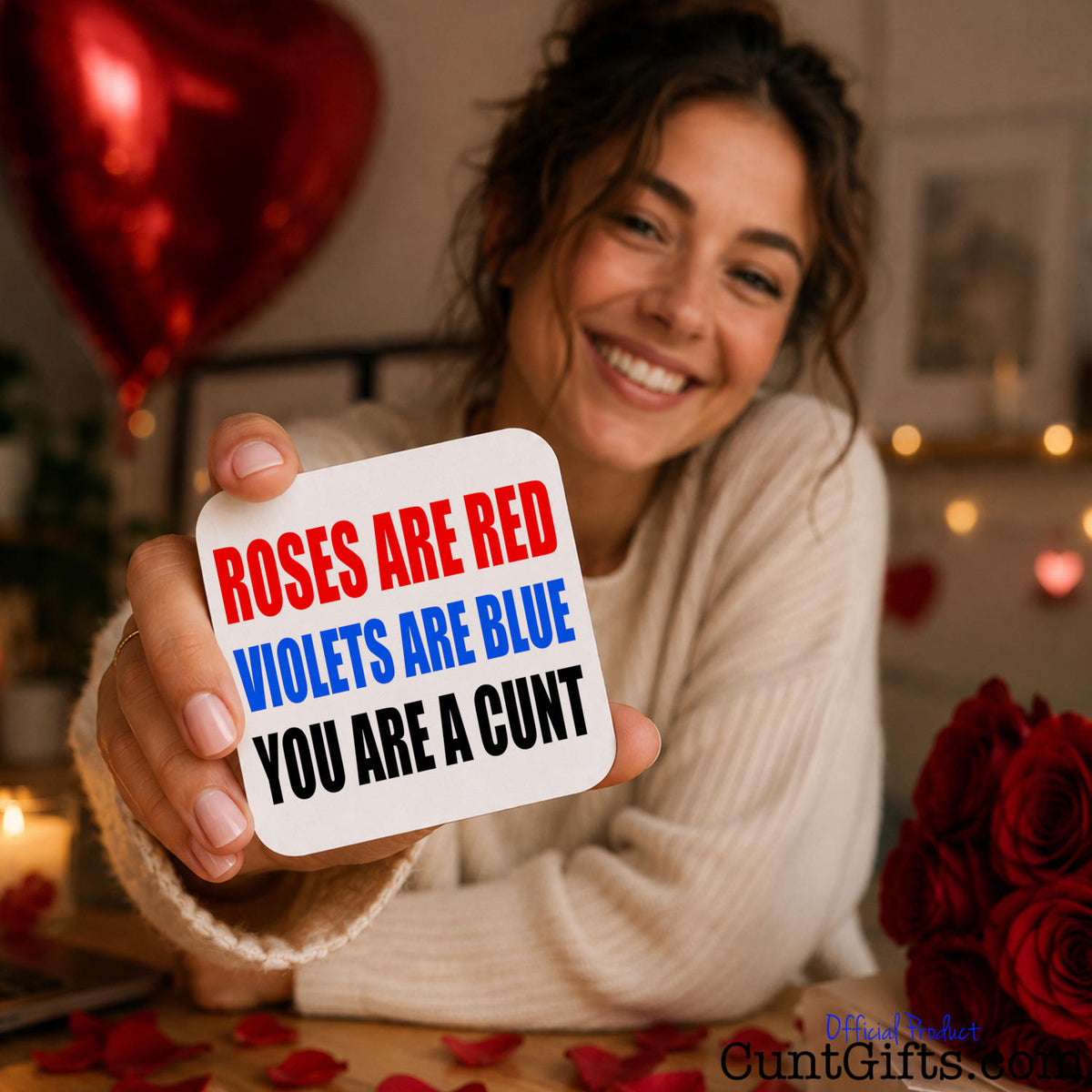 A smiling woman in a cream sweater holds up a white square drinks coaster with the phrase 'ROSES ARE RED VIOLETS ARE BLUE YOU ARE A CUNT' in red, blue, and black text. The background is a cozy indoor setting with a red heart balloon, blurred lights, and red roses.