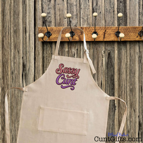 Linen apron with 'Sassy Cunt' text on a wooden wall with hooks.