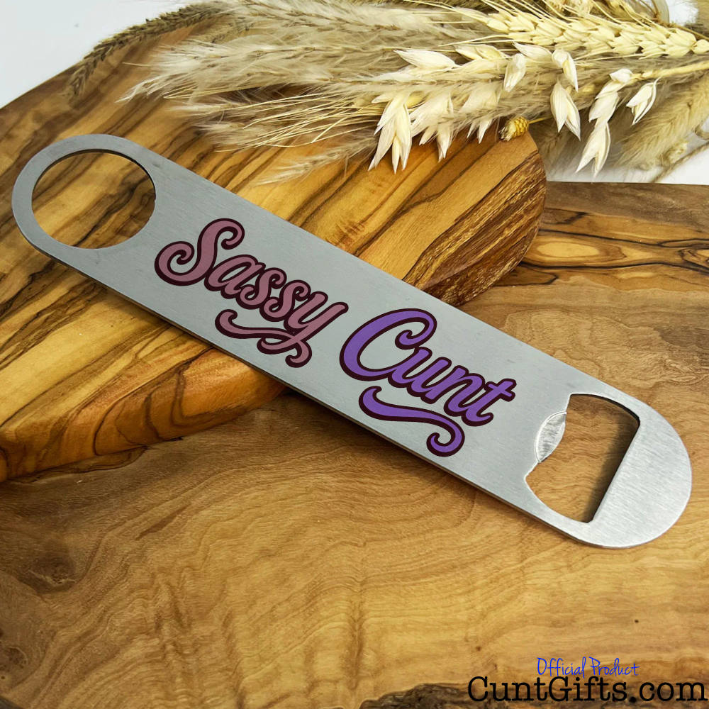 Metal bottle opener with 'Sassy Cunt' text on a wooden board with wheat.