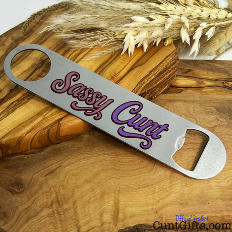 Metal bottle opener with 'Sassy Cunt' text on a wooden board with wheat.