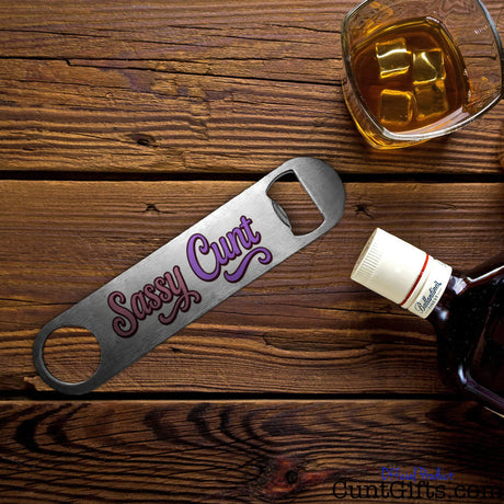 Metal bottle opener with 'Sassy Cunt' text on a wooden surface with a glass of whiskey.