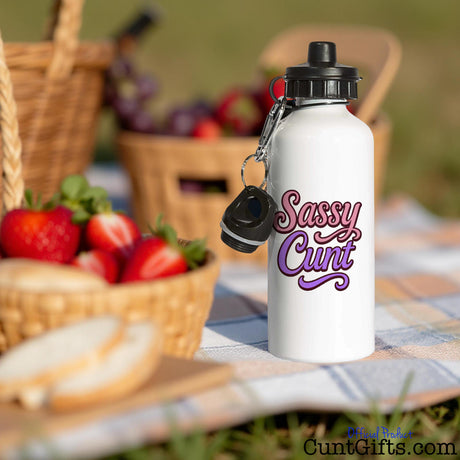 White water bottle with 'Sassy Cunt' text on a picnic setting with strawberries and bread.