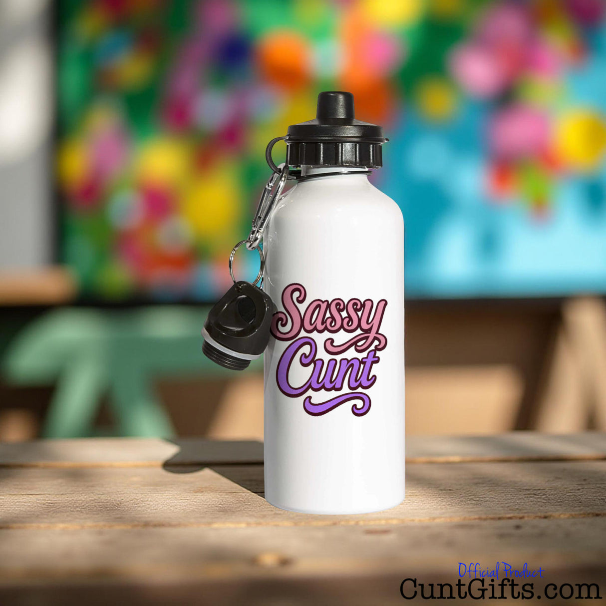 White water bottle with 'Sassy Cunt' text on a wooden table with a colorful abstract painting in the background.
