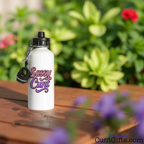 White water bottle with 'Sassy Cunt' text on a wooden surface with plants in the background
