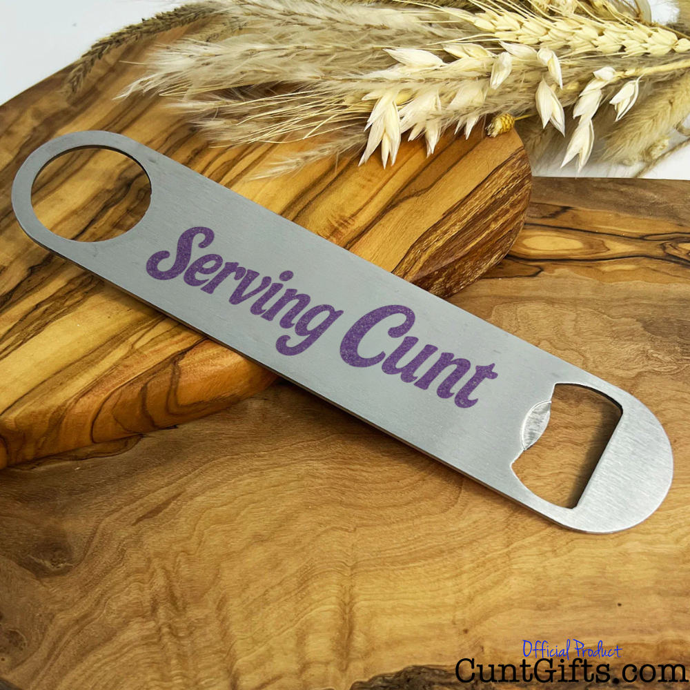 Metal bottle opener with 'Serving Cunt' text on a wooden chopping board with wheat.