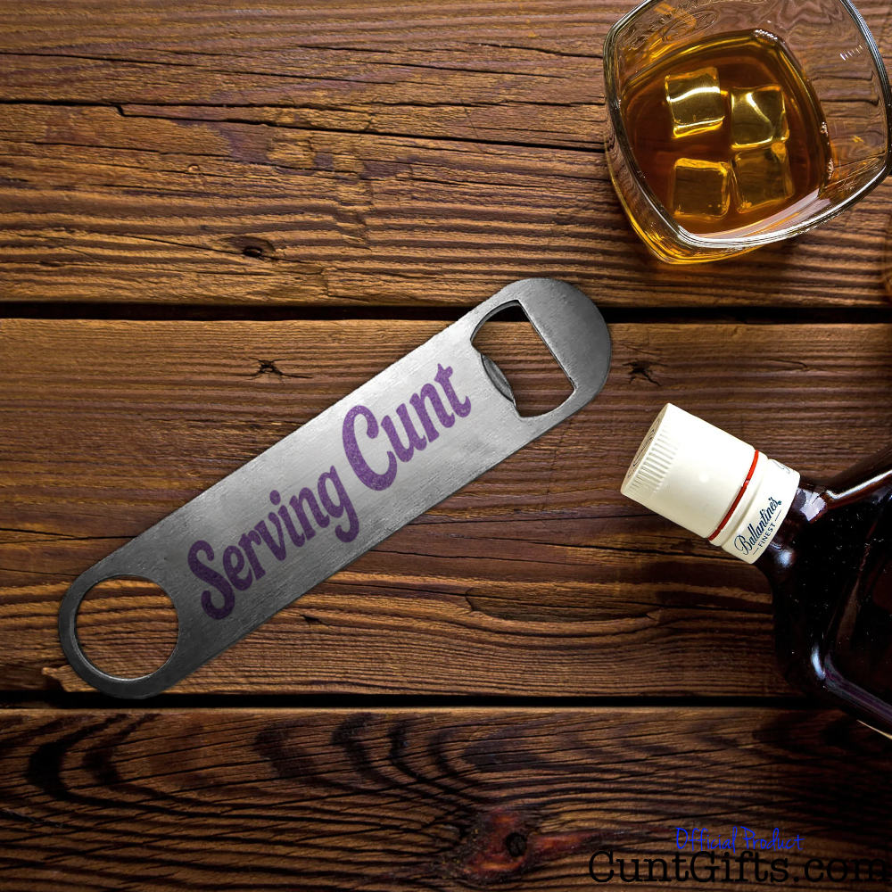 Bottle opener with 'Serving Cunt' design on a wooden surface with a glass of whiskey.