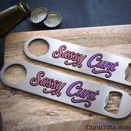 Two metal bottle openers with 'Sassy Cunt' text on a wooden surface.