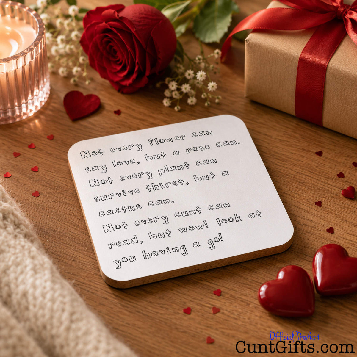 A square white "Not every cunt can read" drinks coaster with a humorous quote printed in gray text, styled on a wooden table with a red rose, candle, gift box, and red hearts.