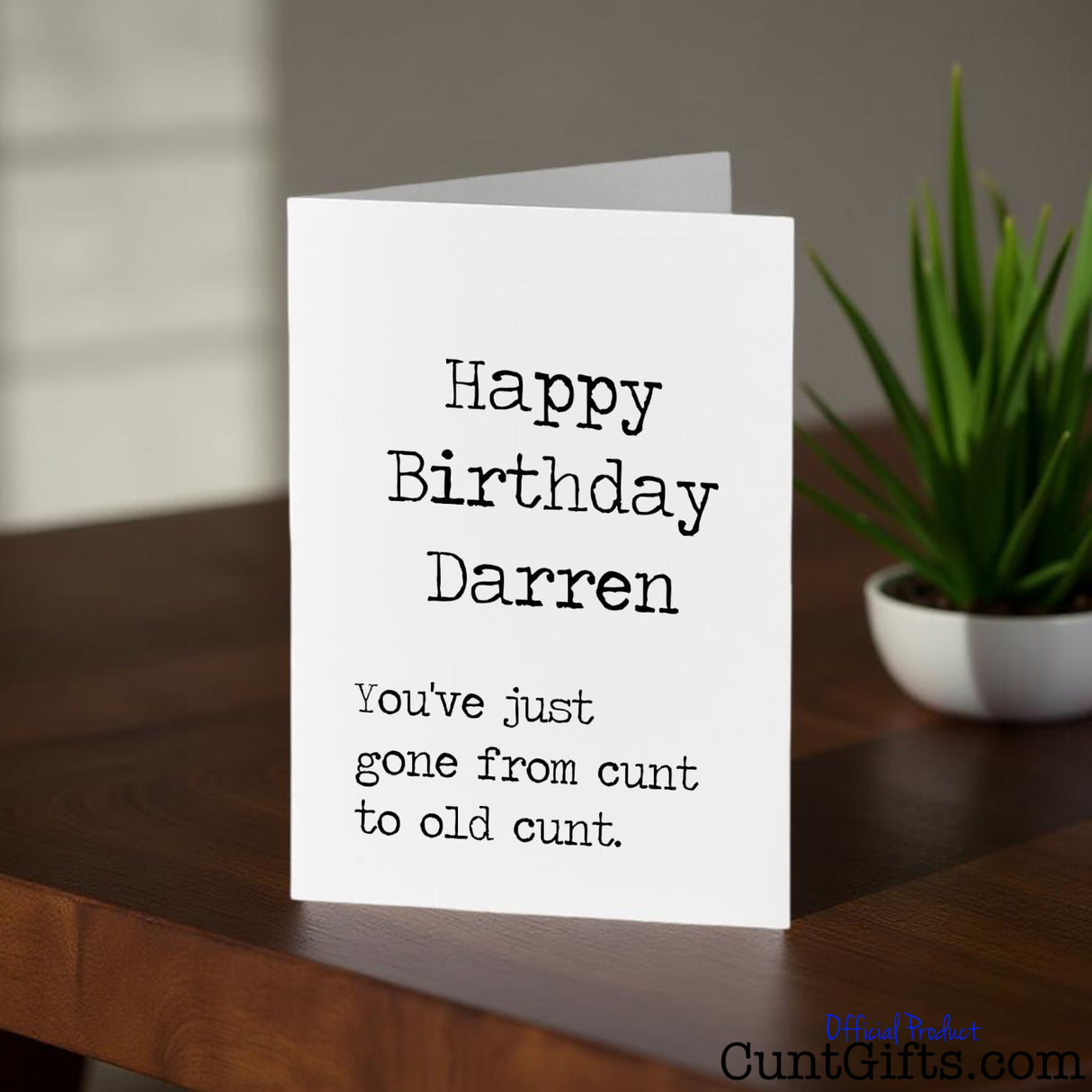 Happy Birthday ANY NAME Old Cunt birthday card on a wooden surface with a plant in the background