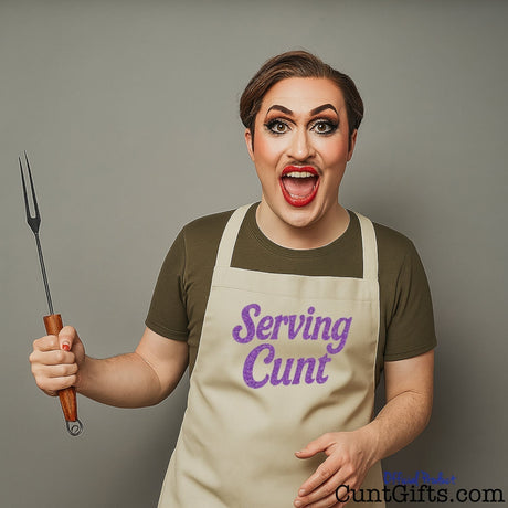 Man in full makeup wearing a “Serving Cunt” apron, holding BBQ tongs and serving fierce energy with a big grin