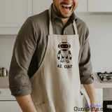 Man wearing a funny AI Cunt apron featuring an orange-eyed cartoon robot design in a kitchen.