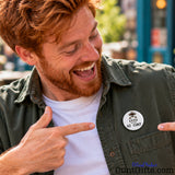 Laughing man pointing at his AI Cunt novelty pin badge while wearing a black jacket over a white T-shirt.