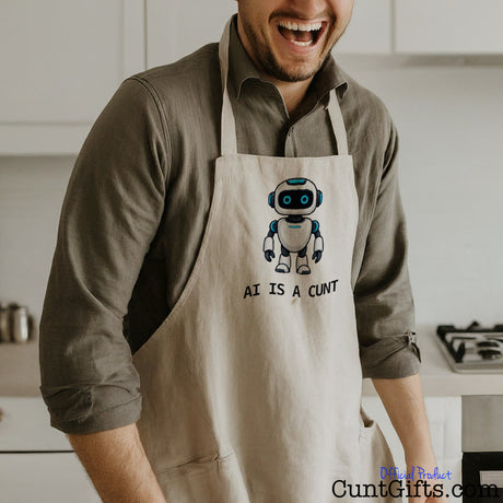 Man wearing a funny AI Is a Cunt apron featuring a cheeky robot design while standing in a kitchen.