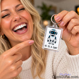 Laughing blonde woman holding up a funny AI Is a Cunt keyring and pointing at its cartoon robot design.