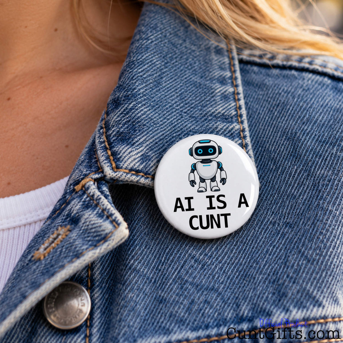 Close-up of a white AI Is a Cunt pin badge featuring a cartoon robot attached to a blue denim jacket.