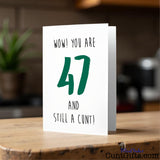 Card with text 'WOW! YOU ARE 47 AND STILL A CUNT!' on a wooden surface.