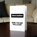 Greeting card with text 'ABRACADABRA! NOPE, YOU ARE STILL A CUNT' on a table in a lounge setting