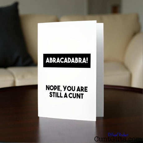 Greeting card with text 'ABRACADABRA! NOPE, YOU ARE STILL A CUNT' on a table in a lounge setting