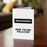 Greeting card with text 'Abracadabra! Nope, you are still a cunt' on a wooden table with a blurred background