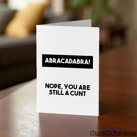 Greeting card with text 'Abracadabra! Nope, you are still a cunt' on a wooden table with a blurred background
