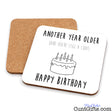 A square white birthday coaster with rounded corners is shown on a white background, with the back of a second cork coaster visible behind it. The front coaster has black text reading “ANOTHER YEAR OLDER (AND YOU’RE STILL A CUNT)” above a simple line drawing of a birthday cake and “HAPPY BIRTHDAY” below.