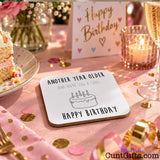 A square white birthday coaster with black text reading 'ANOTHER YEAR OLDER (AND YOU'RE STILL A CUNT)' and 'HAPPY BIRTHDAY' with a simple cake illustration, placed on a pink party table with confetti, a candle, a slice of cake, and a 'Happy Birthday' card in the background.