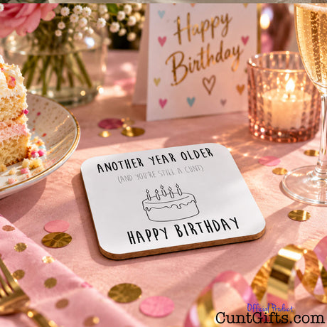 A square white birthday coaster with black text reading 'ANOTHER YEAR OLDER (AND YOU'RE STILL A CUNT)' and 'HAPPY BIRTHDAY' with a simple cake illustration, placed on a pink party table with confetti, a candle, a slice of cake, and a 'Happy Birthday' card in the background.