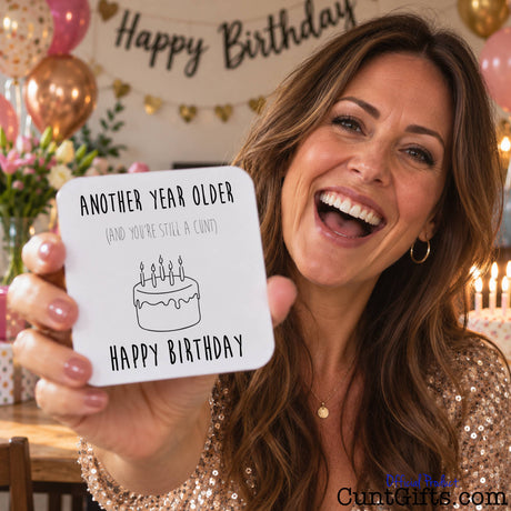 A woman smiling and holding up a square white birthday drinks coaster  that reads 'ANOTHER YEAR OLDER (AND YOU'RE STILL A CUMT)' with a simple cake illustration and 'HAPPY BIRTHDAY' below. The background shows a decorated birthday setting with balloons, a 'Happy Birthday' banner, and candles.
