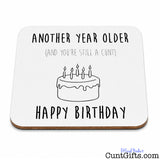 A square white birthdaydrinks coaster  with rounded corners, featuring a black line drawing of a cake with candles and the text 'ANOTHER YEAR OLDER (AND YOU'RE STILL A CUNT)' above and 'HAPPY BIRTHDAY' below.