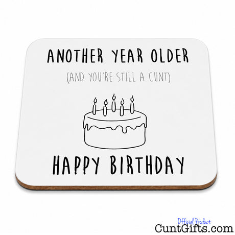 A square white birthdaydrinks coaster  with rounded corners, featuring a black line drawing of a cake with candles and the text 'ANOTHER YEAR OLDER (AND YOU'RE STILL A CUNT)' above and 'HAPPY BIRTHDAY' below.