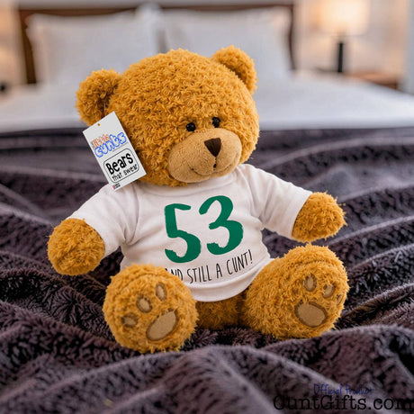 A brown plush personalised teddy bear sitting on a dark textured blanket in a bedroom setting, wearing a white T-shirt with green text that reads '53 AND STILL A CUNT!'. 