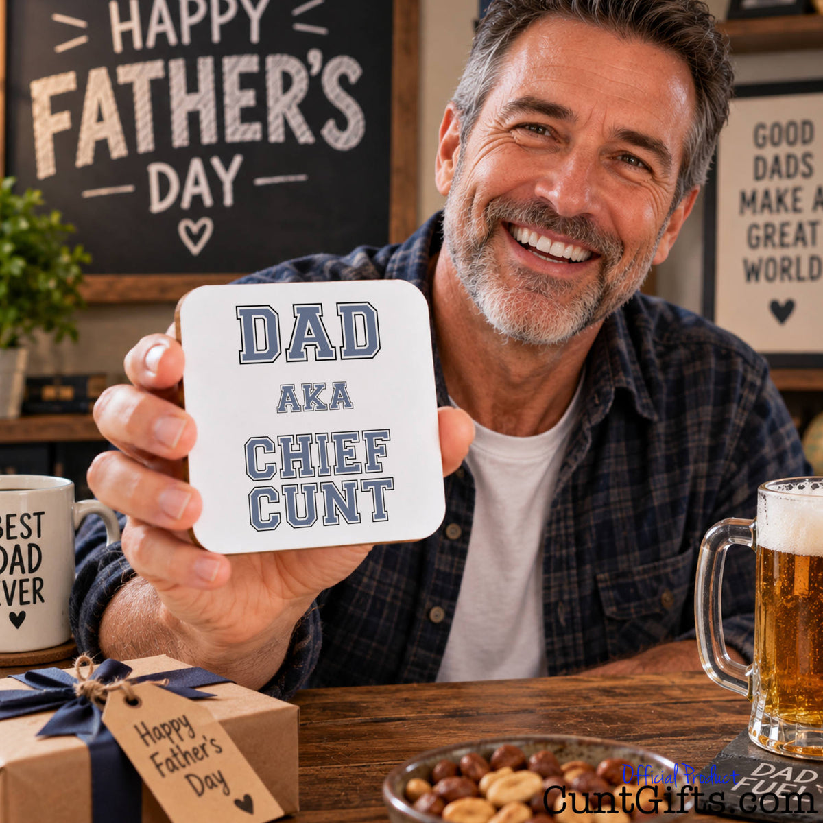 A smiling man in a plaid shirt holds up a square white coaster with the text 'DAD AKA CHIEF CUNT' in grey lettering. The background shows a cozy Father's Day setting with a 'Happy Father's Day' sign, a mug, a beer mug, and a gift box.
