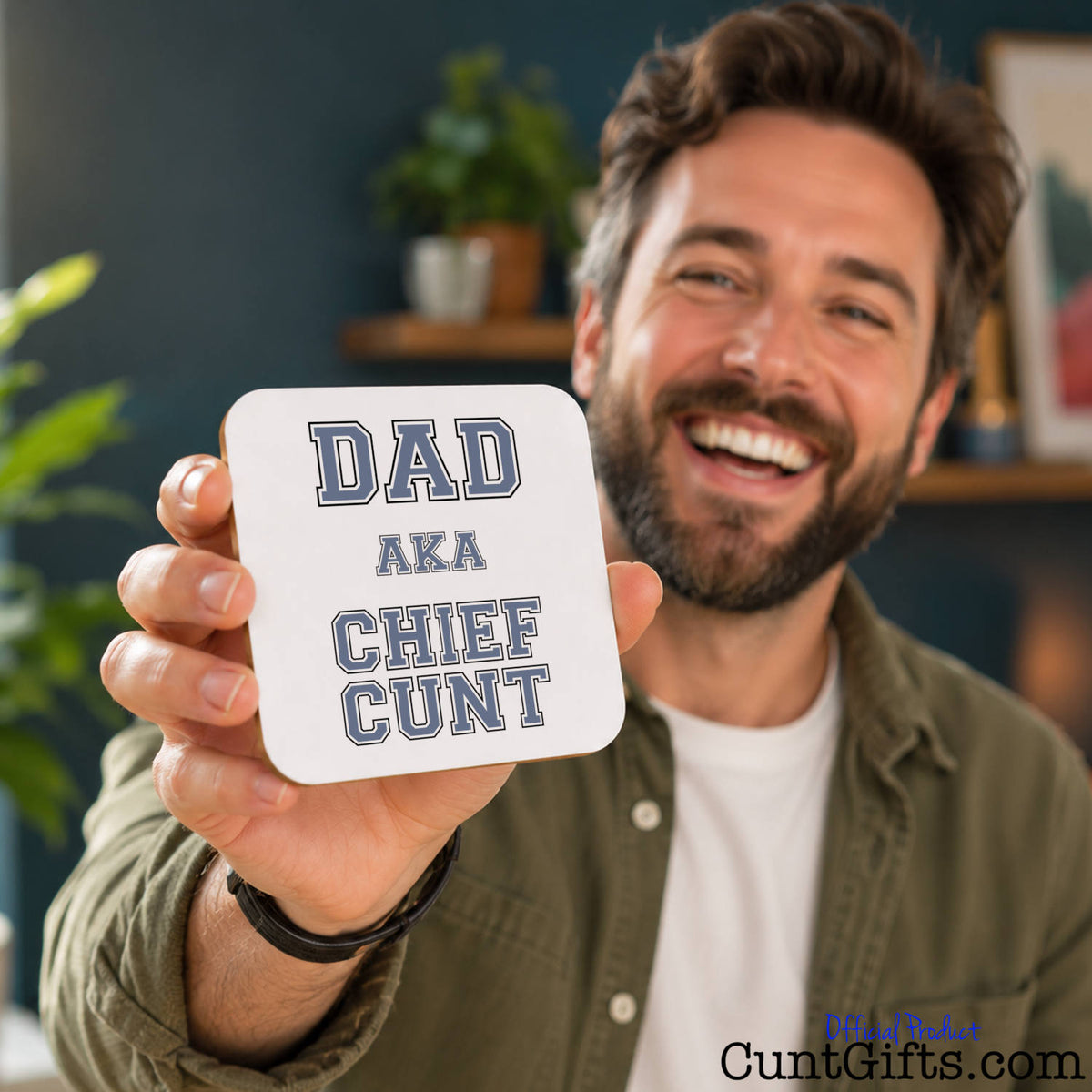 A smiling man holds up a square white coaster with the text 'DAD AKA CHIEF CUNT' in grey lettering. The background shows a cozy indoor setting with a dark blue wall, wooden shelves, and a potted plant.