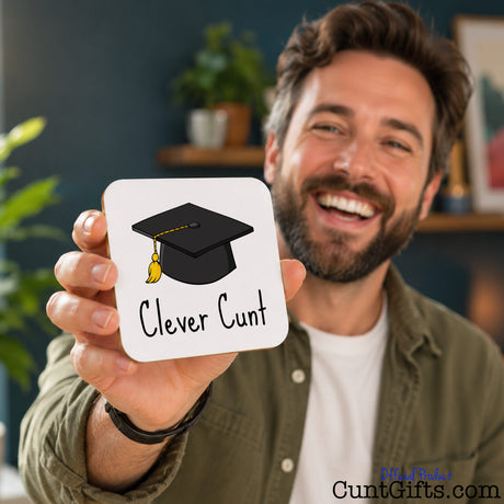 A smiling man holds up a square white drink coaster featuring a black graduation cap illustration and the text 'Clever Cunt'. The background shows a softly blurred indoor setting with a dark blue wall and shelves.