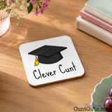 A square white coaster with rounded corners sits on a wooden table, printed with a black graduation cap and the words 'Clever Cunt' in black script. Decorative books, flowers, and a bowl of grapes are visible around it.