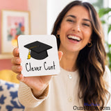 A smiling woman holds up a square white drink coaster featuring a black graduation cap with a yellow tassel and the text 'Clever Cunt' in black. The background shows a softly blurred indoor room with warm tones.