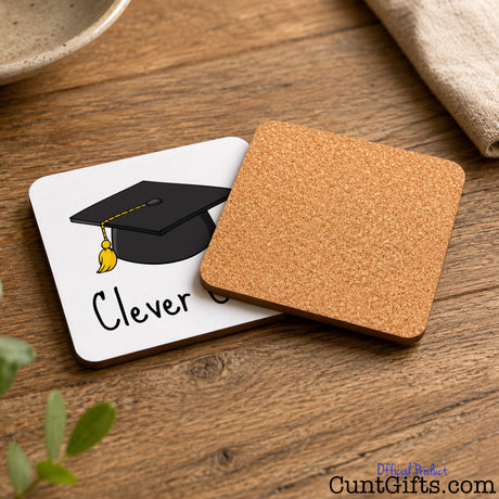 Clever Cunt Drink Coaster on a wooden table showing the reverse side that features a cork backing. They are next to a plant, bowl and tea towel.