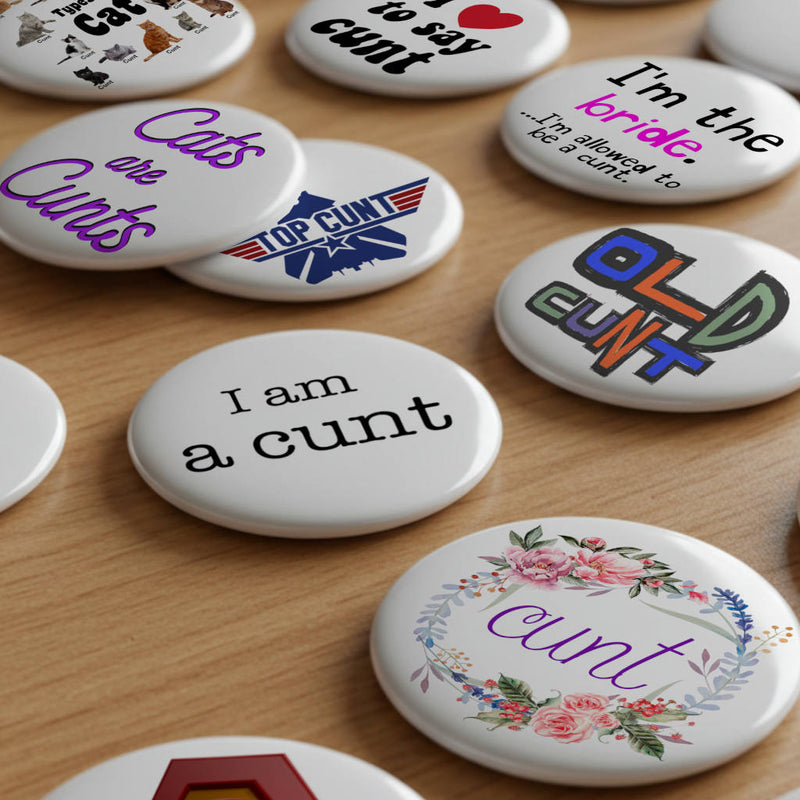 Cunt Badges Collection showing a range of pin badges on a wooden desk all featuring designs with the word cunt.