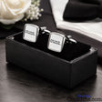 A pair of square silver cufflinks with white faces and the word 'cunt.' printed in black, displayed in a black presentation box on a dark tabletop with a white rose and folded white fabric in the background.