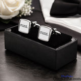 A pair of square silver cufflinks with white faces and the word 'cunt.' printed in black, displayed in a black presentation box on a dark tabletop with a white rose and folded white fabric in the background.