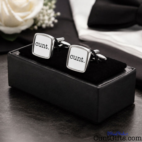 A pair of square silver cufflinks with white faces and the word 'cunt.' printed in black, displayed in a black presentation box on a dark tabletop with a white rose and folded white fabric in the background.