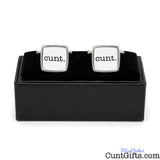 A pair of square silver cufflinks with white faces printed with the word 'cunt.' in black lowercase letters, displayed on a black presentation box against a white background.